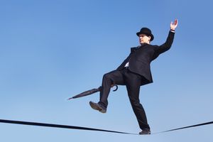 A man in a black suit and bowler hat balances precariously on a tightrope, holding an umbrella in one hand, against a clear blue sky. The image symbolizes navigating challenges with agility and balance in an uncertain environment.